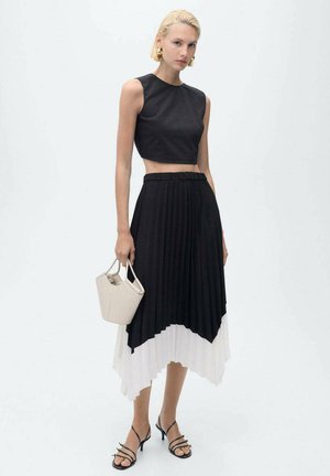 Top corto nero abbinato a una gonna asimmetrica a pieghe bianca e nera. La modella tiene in mano una piccola borsa beige e indossa sandali neri con lacci.