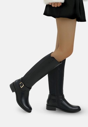 Gambe che indossano stivali di pelle neri al ginocchio con fibbie, abbinati a una gonna corta nera e a una giacca bianca con pelliccia.