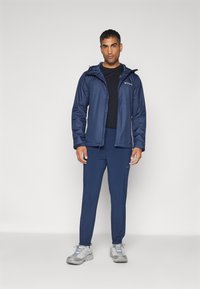 Veste imperméable bleu marine avec capuche, portée par-dessus un haut noir et un pantalon bleu marine. Les caractéristiques incluent des zips et des poignets élastiques. Des baskets grises complètent le look.