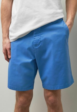 Mann trägt leuchtend blaue Shorts und ein weißes, in die Hose gestecktes T-Shirt, mit einer Hand in der Tasche, stehend vor einem schlichten hellen Hintergrund.
