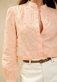 Blusa rosa ricamata con colletto alto, motivi floreali e chiusura a bottoni; abbinata a pantaloni bianchi a vita alta e una cintura marrone.