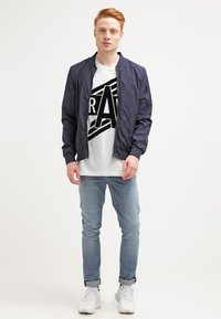 Jachetă de bombardier bleu cu o textură netedă, tricou alb cu grafică și litere negre, blugi deschiși la culoare și adidași albi.