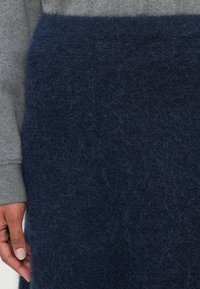 Námorně modrý vlněný svetr s měkkou, chlupatou texturou. Obsahuje šedou látkovou rukáv, který vykazuje vrstvený design. Detailní pohled.