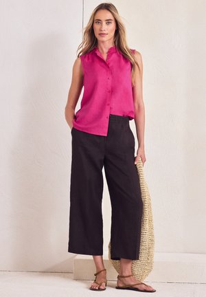 Kvinde iført ærmeløs fuchsia skjorte med knapper, sorte vidde cropped bukser, brune sandaler, der holder en stor flettet taske.