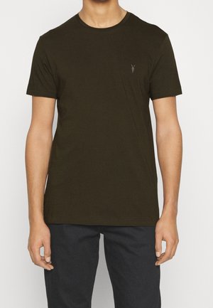 Mężczyzna ubrany w gładki ciemnozielony t-shirt z krótkim rękawem i czarne spodnie, stojący z rozluźnionymi ramionami wzdłuż ciała.