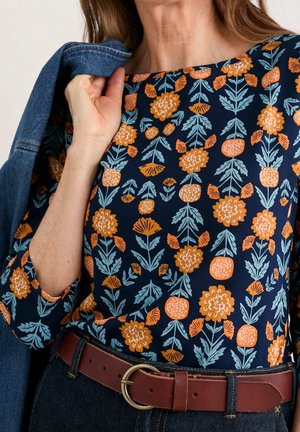 Vrouw draagt marineblauwe top met oranje en lichtblauw bloemenpatroon, houdt spijkerjack over schouder, bruine riem met ronde gesp zichtbaar.