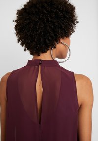 Blouse bordeaux avec un panneau arrière transparent, col montant et fermeture à boutons, présentant un détail en trou de serrure. Grande boucle d'oreille en argent visible.