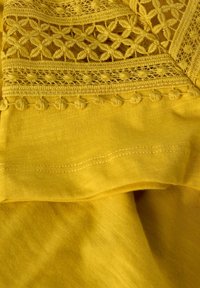 Tela amarilla con intrincados detalles de encaje y bordado. Textura suave, presenta costuras decorativas y lazos acentuados a lo largo del borde.