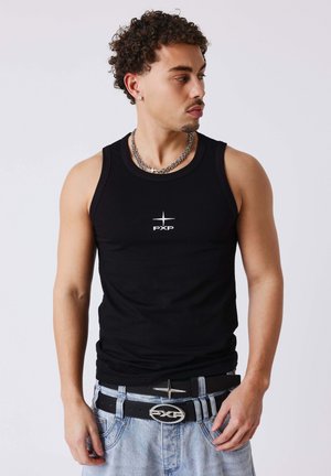 Jeune homme aux cheveux bouclés portant un débardeur noir avec le logo "PXP", un collier chaîne argenté et un jean bleu clair avec une ceinture noire.