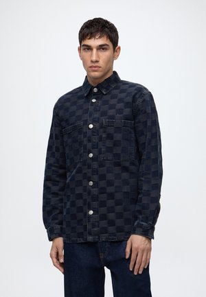 Jeune homme portant une chemise à carreaux bleu foncé avec boutons et deux poches poitrine, ainsi qu'un jean bleu foncé, debout devant un fond uni.