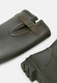 Tumed rohelised kummisaapad, millel on tekstuuriga pind, buckliga kaunistus, tugevdatud konts ja kindel mustriga tall.