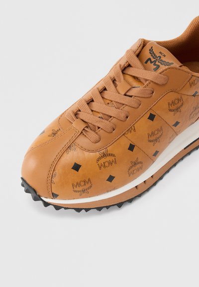 Αθλητικό παπούτσι από δέρμα ταμπά με επιφάνεια διακοσμημένη με μαύρα λογότυπα MCM και διαμάντι σχέδια. Στρογγυλερή μύτη, καφέ κορδόνια και λευκή ελαστική σόλα.
