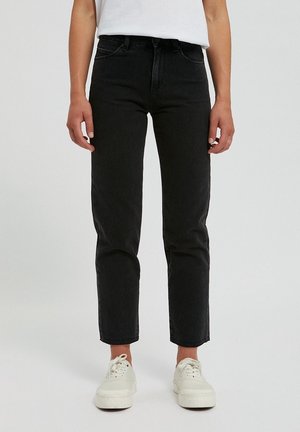 Person wearing black straight-leg jeans, white sneakers, and a white shirt, standing against a plain light background.