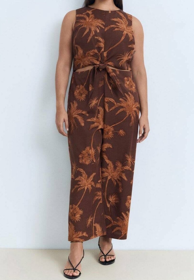 Femme portant une combinaison marron sans manches avec des imprimés de palmiers orange et un détail noué à l'avant. Sandales noires à brides aux pieds.