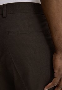 Dunkelbraune Hose mit strukturiertem Stoff, schmaler Passform, Bund mit Gürtelschlaufen und einer einzelnen Gesäßtasche mit verdeckter Öffnung.