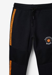 Zwarte trainingsbroek met oranje zijkanten, logo "Basketball Hero" en gaasaccenten op de knieën. Elastische tailleband en zachte textuur.
