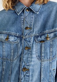 Helehall sinist värvi teksajope, millel on kraega kaelus, kuldsed nupud, kaks rinnataskut ja nähtavad õmblusdetailid.