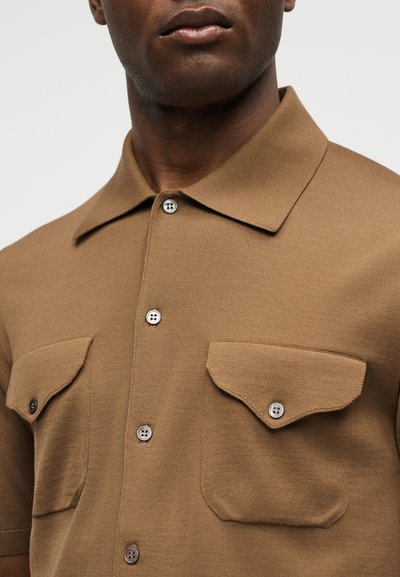 Άνδρας που φοράει καφέ κοντομάνικο πουκάμισο με γιακά και δύο κουμπωτές τσέπες στο στήθος, σε απλό φόντο.