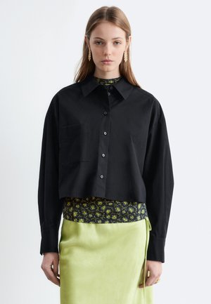 Mladá žena oblečená v čiernej krátkej košeli na gombíky cez kvetovaný top a limetkovo zelenú sukňu, stojaca pred jednoduchým bielym pozadím.