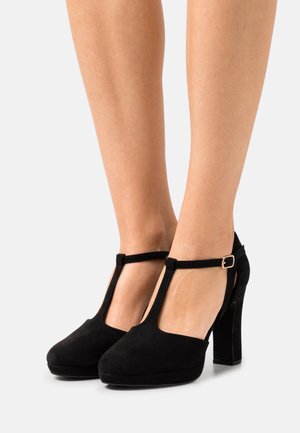 Escarpins T-strap en suede noir avec un bout arrondi, une finition texturée et un talon haut carré accentué par une petite boucle métallique.
