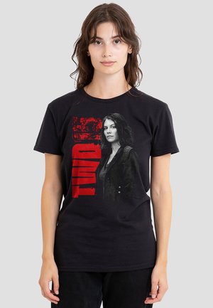 Young woman wearing black T-shirt featuring grayscale portrait of a serious woman and bold red text "TWD" on plain background.