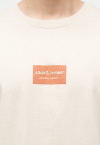 Κοντινό πλάνο ενός κρεμ T-shirt με ένα κεντρικά τοποθετημένο πορτοκαλί ορθογώνιο λογότυπο που γράφει "Jack & Jones Definition of Denim."