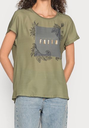 Frau trägt ein olivgrünes T-Shirt mit einem grauen Quadrat-Grafik und dem Wort "FAITH" in goldenen Buchstaben, kombiniert mit hellblauen Jeans.
