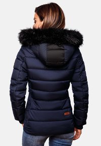Marineblå pufferjakke med en aftagelig sort faux fur-hood, ribbede manchetter og et quiltet design. Har et brunt logo-patch på siden.