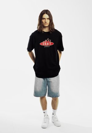 Camiseta negra de manga corta con logo de llamas rojas y texto "GRMY", combinada con pantalones cortos de mezclilla azul claro y zapatillas blancas. Fondo simple.