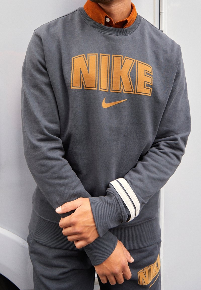 Grijze sweatshirt met oranje "NIKE" tekst en logo. Bevat een geribbelde ronde hals, gestreepte accenten op de manchetten en een ontspannen pasvorm.