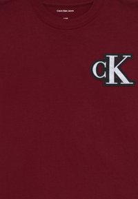 Burgundy T-shirt made of cotton with a round neckline, featuring an embroidered black and white 'cK' logo on the left chest area.