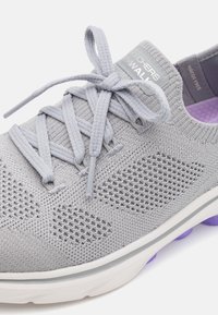 Chaussure de sport gris clair avec une tige en maille respirante, des motifs texturés, des lacets plats, et une semelle intermédiaire blanche et violette.