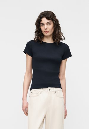 Femme aux cheveux bouclés mi-longs portant un haut noir ajusté à manches courtes et un pantalon taille haute blanc cassé, sur un fond uni.