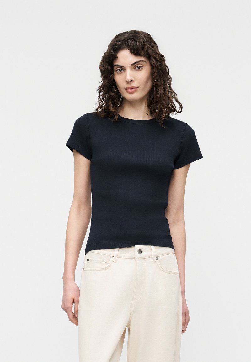 Mujer con cabello rizado hasta los hombros, vistiendo una camiseta negra ajustada de manga corta y pantalones de talle alto color blanco roto, frente a un fondo sencillo.