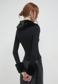 Eine rothaarige Frau, die von hinten zu sehen ist, trägt ein schwarzes, geripptes Langarmoberteil mit Kunstfellkragen und -manschetten sowie einen schwarzen Rock. Ihre Hände sind hinter dem Rücken verschränkt.