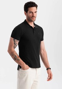 Muž s tetováním oblečený v upnutém černém polo tričku a světlých béžových kalhotách, stojící sebevědomě na neutrálním pozadí.