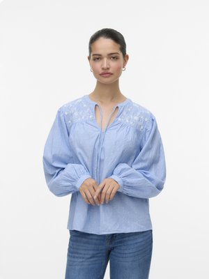 Light blue blouse with embroidered floral details, relaxed fit, long puff sleeves, and a tie neckline. Paired with medium-wash jeans.