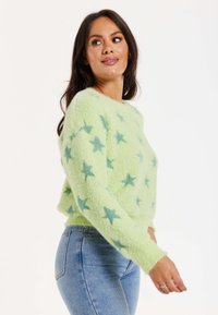 Green fuzzy sweater with blue star patterns, round neck, long sleeves, and ribbed hem, paired with light blue denim jeans.