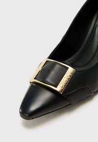 Pantofii din piele neagră, cu vârful ascuțit, prezintă o cataramă mare din aur cu "DKNY" gravat, o suprafață texturată și un design al tocului curbat.