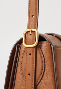 Borsa a mano in pelle marrone con una texture liscia, accessori in metallo dorato e una fibbia rettangolare sulla tracolla. Presenta cuciture e dettagli precisi.