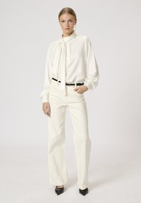 Weißes Blusenshirt mit Halsband und voluminösen Ärmeln, kombiniert mit cremefarbenen weiten Hosen. Das Outfit umfasst einen schwarzen Gürtel und spitze schwarze High Heels.
