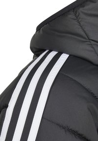 Polstret svart jakke med hette, med tre hvite horisontale striper på skuldrene. Materialet ser glatt og quiltet ut.