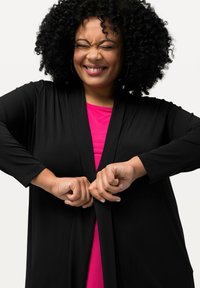 Schwarze, langärmlige Strickjacke, die über einem pinken Top getragen wird. Die Jacke hat einen lässigen Schnitt mit offenem Vorderteil und wird mit beiden Händen geschlossen.