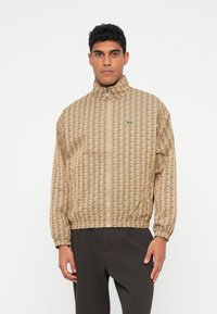 Beige vindjakke med geometrisk print, høj krave, fuld lynlås og elastiske ærmer. Har grønt logo på venstre bryst.