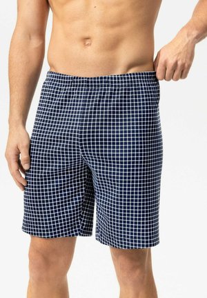 Hemdloser Mann trägt marineblaue und weiße karierte Shorts und zieht mit einer Hand leicht am Bund.