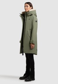 Olivovo zelená dlhá parka s kožušinou podšívkou na kapucni, predným zipsom a bočnými vreckami; obsahuje čierne kovanie a nášivku s logom na rukáve.
