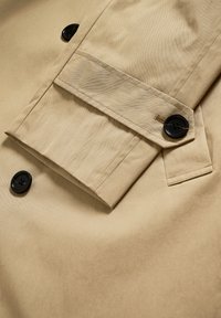 Bézs színű ballonkabát anyag sima textúrával, fekete gombbal és részletes varrással a mandzsettán és a hajtókán.