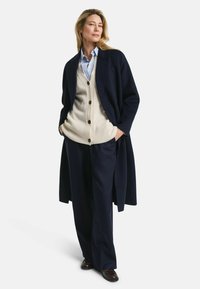 Navy wool coat over beige knit cardigan with buttons, paired with dark pants and brown shoes. Smooth textures; tailored design.