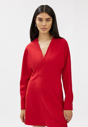 Punainen mekko, jossa on pitkät hihat ja V-pääntie. Valmistettu sileästä kankaasta, jossa on istuva muotoilu saumoilla ja yksinkertainen siluetti.