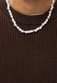 Valkoinen helminauha hopeisilla yksityiskohdilla, esillä ruskean neulepaidan päällä. Pyöreä muoto, erikokoisia helmiä, kiiltävä pinta.